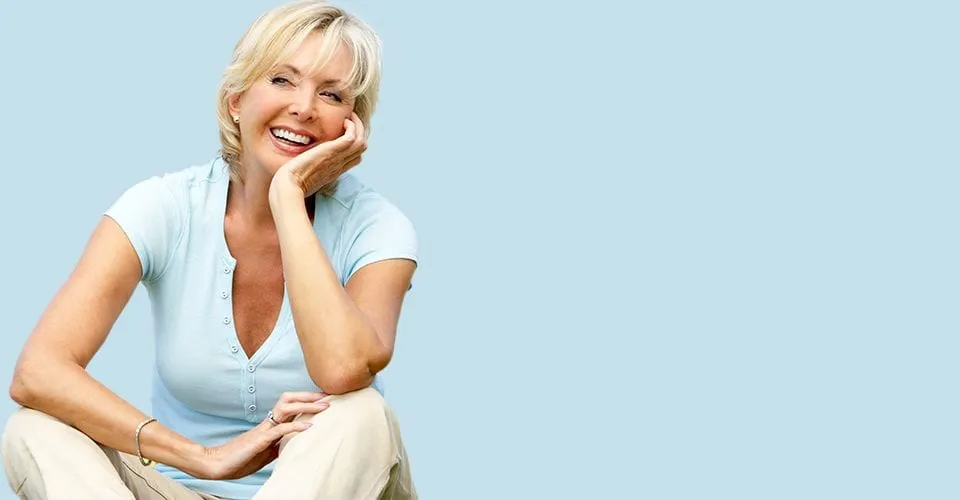 micro_needling_big_2 Mature woman wearing a light blue v-cut t-shirt is smiling and resting her elbows on her knees.