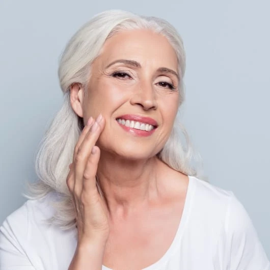 Smiling mature woman touching her facial skin with her fingers in front of gray background.